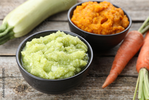 Tasty puree in bowls, zucchini and carrots on wooden table, closeup photo