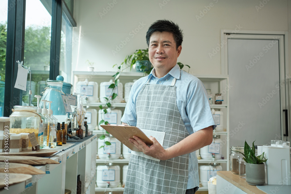 Portrait of an Asian male shopkeeper smiling and looking at camera, arranges natural products at refill store, zero-waste grocery, and plastic-free, eco environment-friendly, sustainable lifestyles. Portrait of an Asian male shopkeeper smiling and looking at camera, arranges natural products at refill store, zero-waste grocery, and plastic-free, eco environment-friendly, sustainable lifestyles.