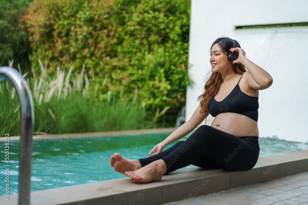 pregnant woman sitting on edge of swimming pool while listening to music with headphone
