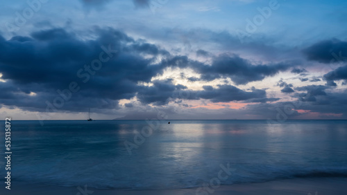 Evening twilight on a tropical island. The turquoise ocean is calm. Blue clouds in the sky  highlighted in pink. Reflection on the water. Foam of waves on a sandy beach. Long exposure. Seychelles. 
