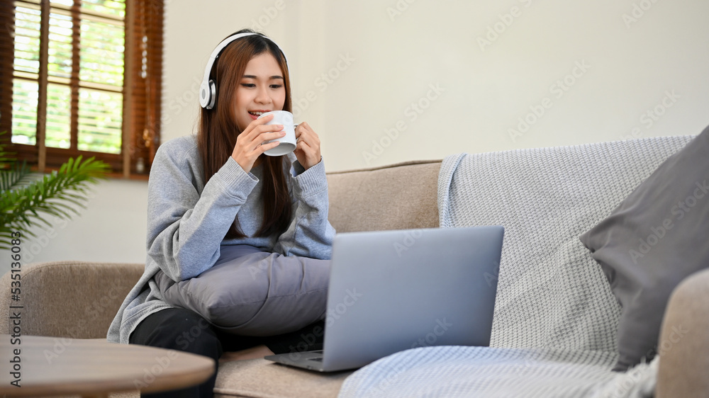 Relaxed Asian female with headphones watching movie on her laptop while sipping coffee Relaxed Asian female with headphones watching movie on her laptop while sipping coffee