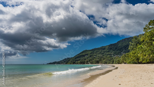 Ocean waves roll onto the beach and foam on the sand. Splashes in the air. A green hill against a blue sky. Picturesque cumulus clouds. Seychelles. Mahe. Beau Vallon