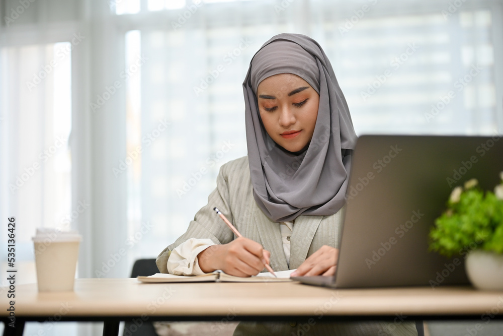 Professional Asian Muslim businesswoman concentrating, writing her plan on a book at her desk. Professional Asian Muslim businesswoman concentrating, writing her plan on a book at her desk.