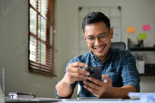 Handsome and happy Asian businessman using his mobile phone at his desk.