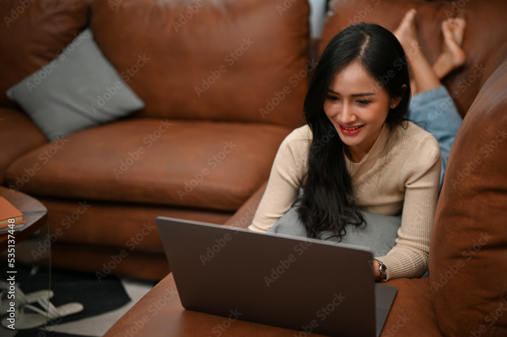 Beautiful Asian female lying on her sofa, using her laptop, reading web blogs, watching movies Beautiful Asian female lying on her sofa, using her laptop, reading web blogs, watching movies