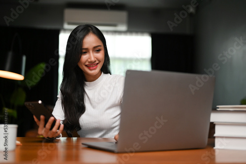 Gorgeous Asian female at her working desk at home, looking at laptop screen, using laptop