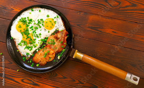 Home made fried eggs and bacon on frying pan with green onion and ground pepper. On wooden table top. Flat lay.