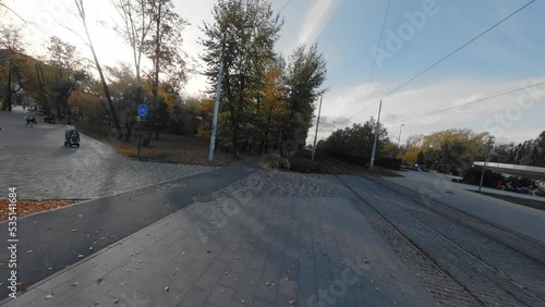FPV drone view flies next to the tram rails on the bike path. Near the park, square, Buildings and trees. People walking in the park. The leaves on the trees are orange, yellow and red. There are many