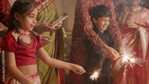 Ethnic Indian Hindu happy smiling cute young boy and a girl or kids in traditional clothes playing with firecrackers or sparklers with the help of family elders or parents during Diwali festival.