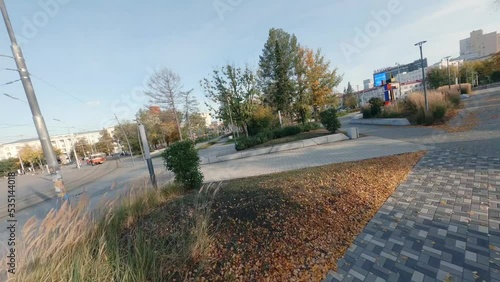 FPV drone view of autumn city park next to the road with cars, house of culture and buildings in city. People are walking. Tram passing by. Autumn sunny day
