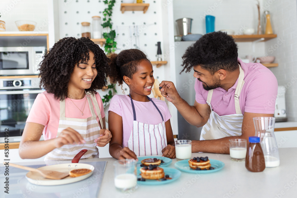 Preparing home bakery. Loving friendly afro american family spouses with daughter cooking food together at kitchen table, making homemade pancakes Preparing home bakery. Loving friendly afro american family spouses with daughter cooking food together at kitchen table, making homemade pancakes
