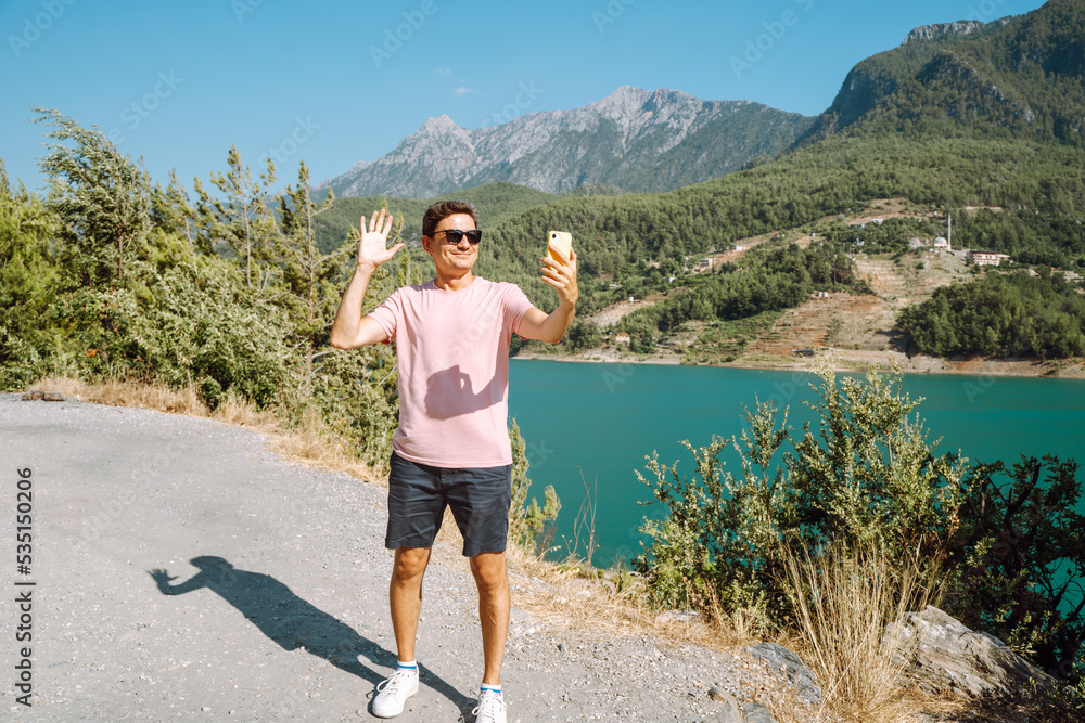 Man taking selfie on mobile phone of mountains lake background. Traveler male having video chat on cellphone on the blue lake outdoors travel adventure vacation.