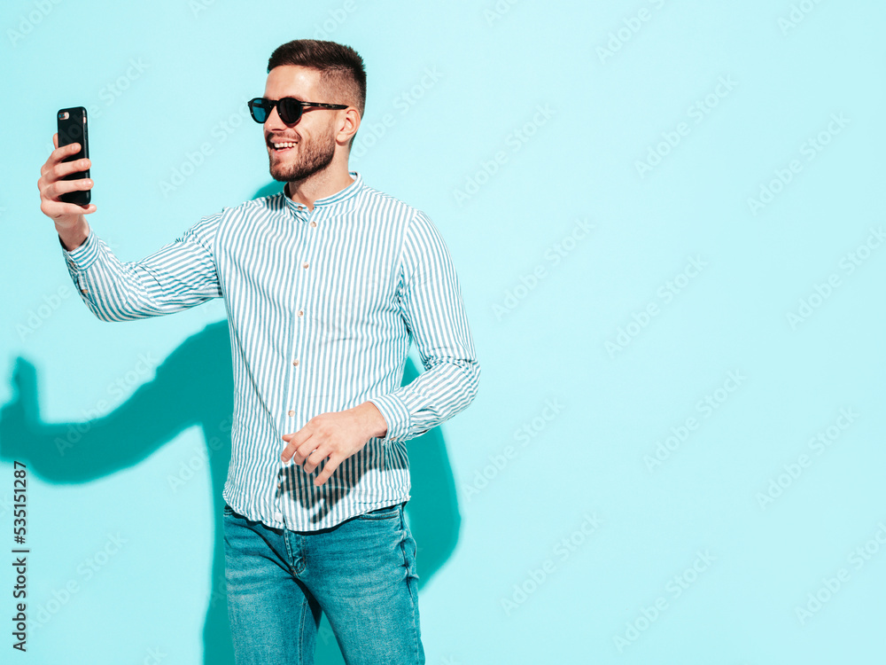 Handsome model. Sexy stylish man dressed in shirt and jeans. Fashion hipster male posing near blue wall in studio. Holding smartphone. Looking at cellphone screen. Using apps. In sunglasses Handsome model. Sexy stylish man dressed in shirt and jeans. Fashion hipster male posing near blue wall in studio. Holding smartphone. Looking at cellphone screen. Using apps. In sunglasses