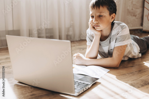 Teenager boy in lying on floor with laptop, having video call and online lesson at home. Schoolboy studying homework. Distance education, remote training concept.