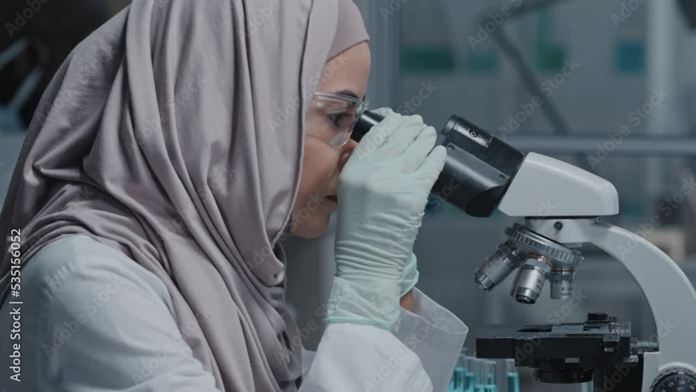Side-view of young Muslim woman in goggles sitting at desk in laboratory, using microscope Side-view of young Muslim woman in goggles sitting at desk in laboratory, using microscope