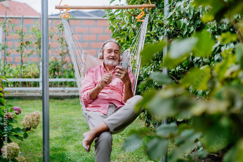Happy senior man sitting in outdoor garden swing, listening music in headphones and scrolling smartphone.