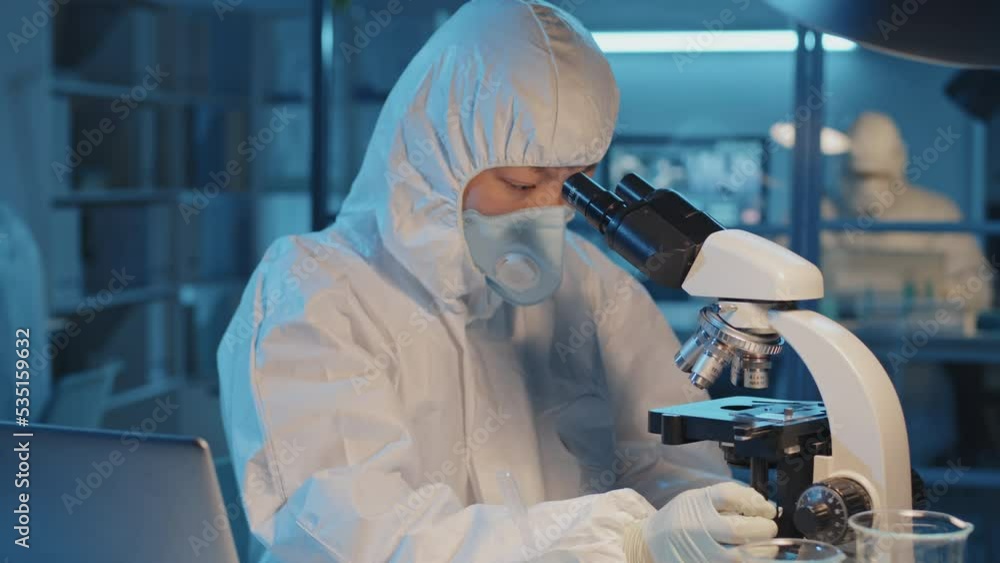 Waist-up of Asian scientist in white hazmat suit and respirator sitting at desk in laboratory, working with hazardous chemicals, using microscope Waist-up of Asian scientist in white hazmat suit and respirator sitting at desk in laboratory, working with hazardous chemicals, using microscope