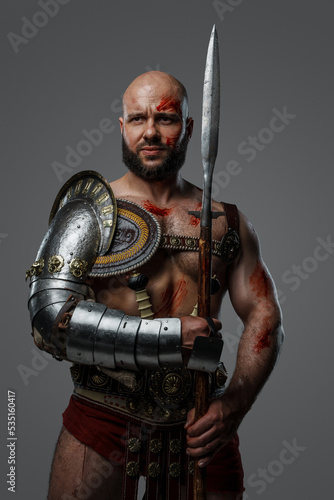Portrait of handsome gladiator with muscular build holding long spear isolated on gray.