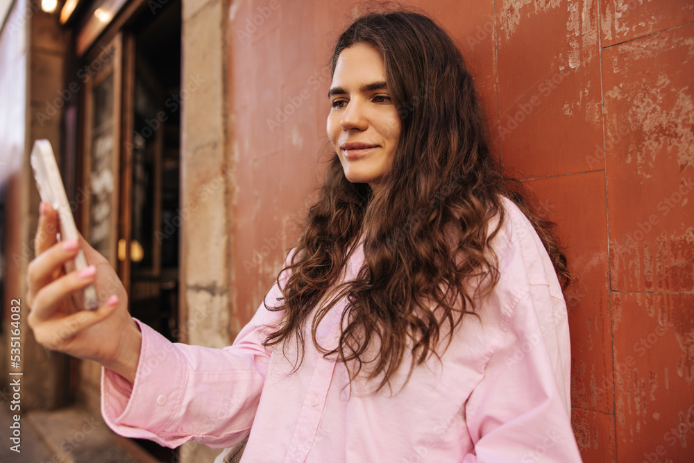 Fototapeta premium Pretty young caucasian girl with wavy hair spends time outdoor and uses her phone. Brunette wears casual pink shirt. Concept selfie, lifestyle.