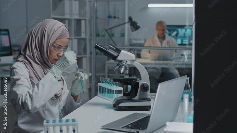 Side view of young female Muslim lab assistant in protective goggles and gloves looking at blue liquid in test tube, sitting at desk in laboratory Side view of young female Muslim lab assistant in protective goggles and gloves looking at blue liquid in test tube, sitting at desk in laboratory