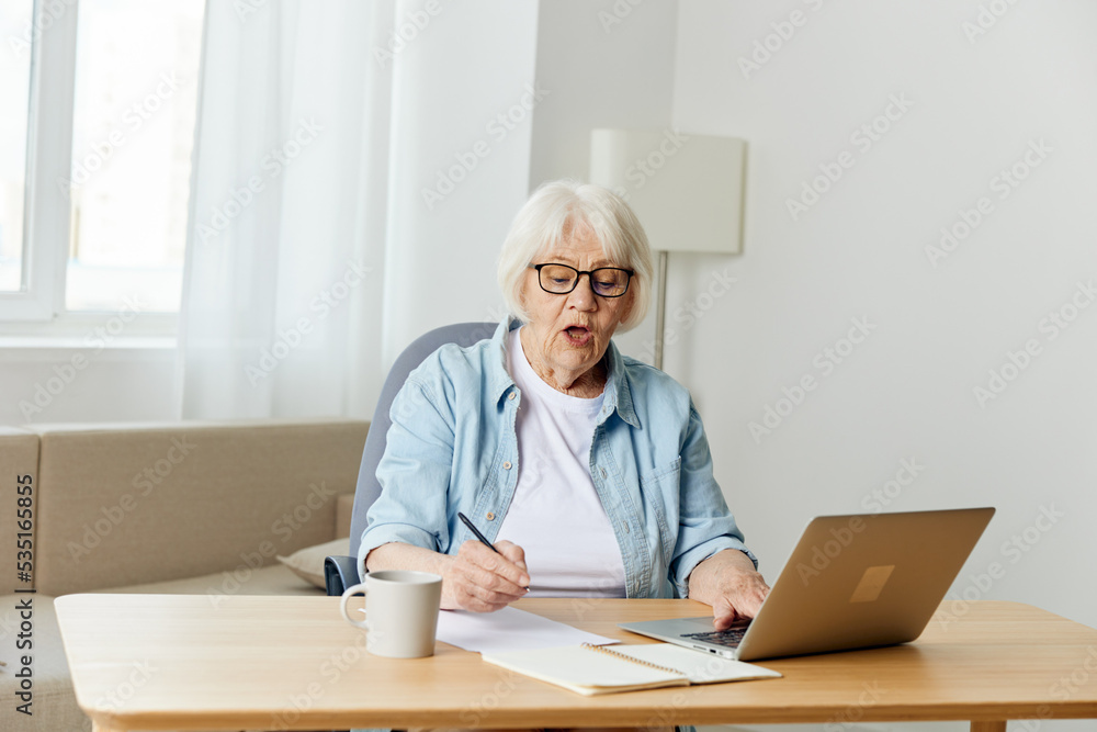 a happy, successful elderly woman is sitting at her desk at home, stylishly dressed in black glasses and happily looking into the camera with a laptop and writing papers on the table a happy, successful elderly woman is sitting at her desk at home, stylishly dressed in black glasses and happily looking into the camera with a laptop and writing papers on the table