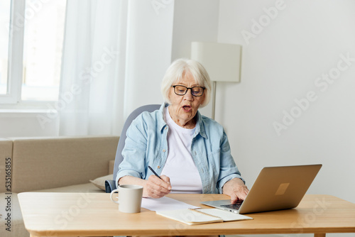 a happy, successful elderly woman is sitting at her desk at home, stylishly dressed in black glasses and happily looking into the camera with a laptop and writing papers on the table
