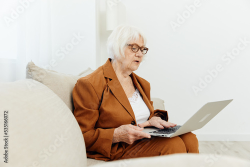an attentive, pleasant elderly woman is sitting in a bright room at home on a cozy sofa and is holding a video conference for company employees © Tatiana