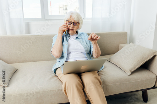 a cute elderly woman is sitting at home working on a laptop resting on the couch and talking on the phone actively gesturing with her hand © Tatiana