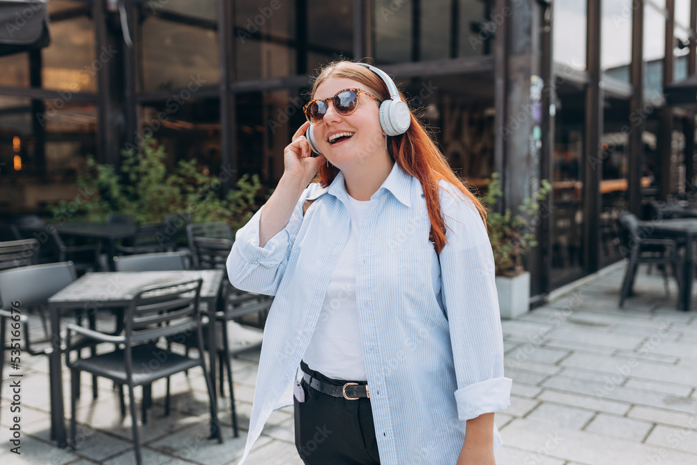 Obraz premium Lovely happy young woman in sunglasess listening to music through wireless earphones on urban background. Music lover enjoying music. Lifestyle concept. Girl walking on city street