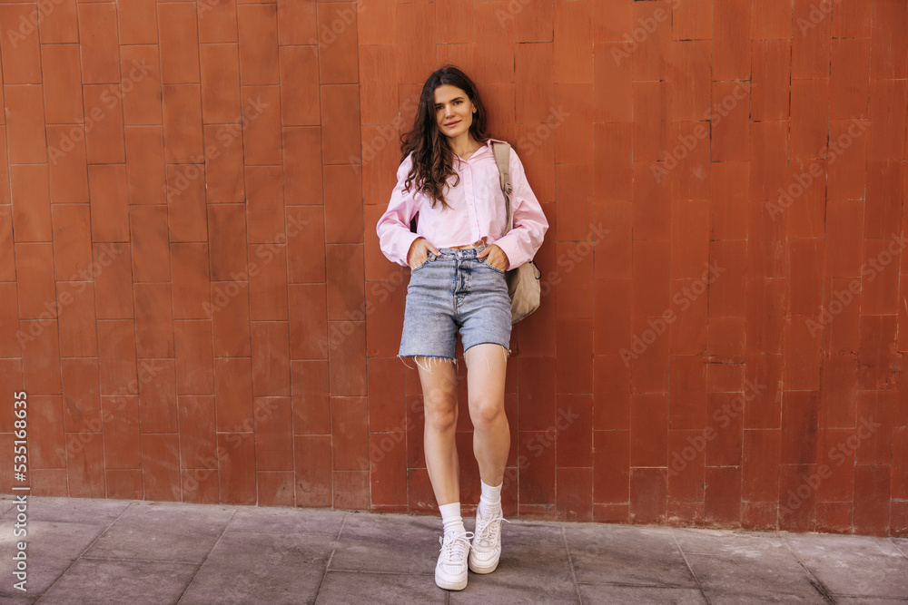 Full length pretty young caucasian woman looking at camera stands near orange brick wall outside. Brunette with wavy hair wears shirt, jeans shirt, shoe and backpack. Concept city life, lifestyle. Full length pretty young caucasian woman looking at camera stands near orange brick wall outside. Brunette with wavy hair wears shirt, jeans shirt, shoe and backpack. Concept city life, lifestyle.