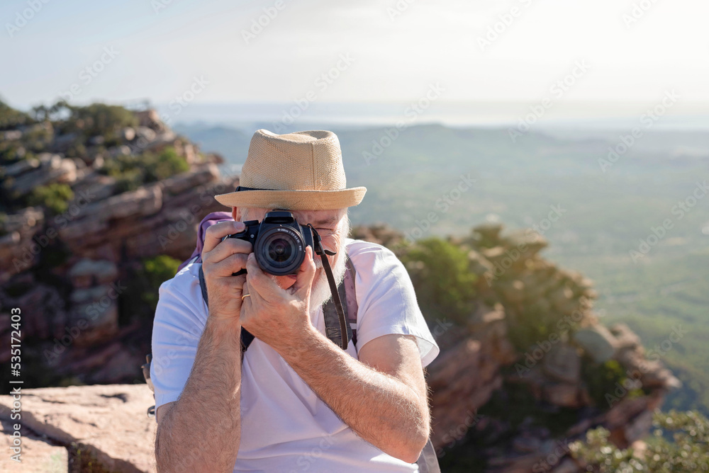 Obraz premium front view of an older hiker sitting on the summit taking a picture with his camera.