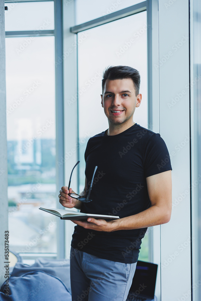 A student manager stands near a large window and reads notes in a notebook. A young man in a black T-shirt with glasses stands in the office near the window A student manager stands near a large window and reads notes in a notebook. A young man in a black T-shirt with glasses stands in the office near the window