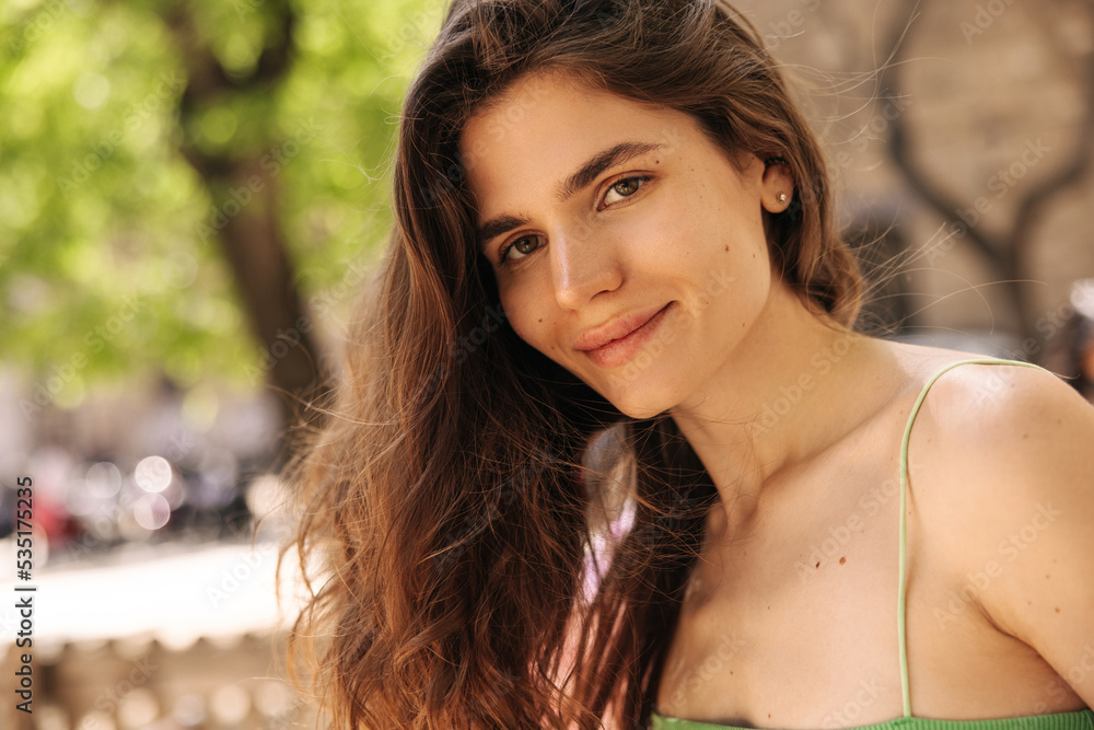 Naklejka premium Close-up pretty young caucasian woman looking at camera with good mood outdoor. Brown wavy hair girl wears green singlet. Concept great mood, solo weekend.