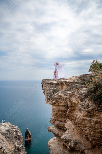 Blonde with long hair on a sunny seashore in a white flowing dress, rear view, silk fabric waving in the wind. Against the backdrop of the blue sky and mountains on the seashore. © svetograph