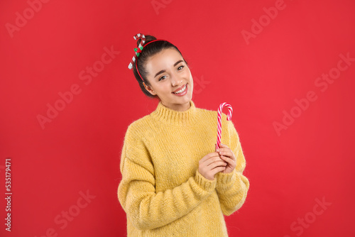 Young woman in yellow sweater and festive headband holding candy cane on red background