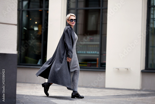Elegant Mature woman walks city street , wears stylish clothes, gray wool coat, long dress, black shoes and glasses. Trending outfit photo