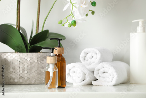 Bottles with dispenser caps  houseplant and towels on white table in bathroom