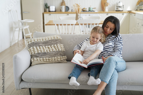 Mom and adopted preschool daughter read book together, sitting on sofa at home. Motherhood, adoption