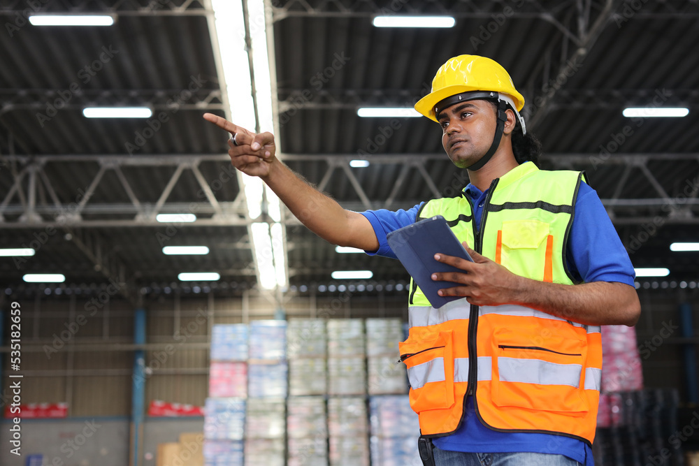 Warehouse workers man with hardhats and reflective jackets using tablet, walkie talkie radio and cardboard while controlling stock and inventory in retail warehouse logistics, distribution center Warehouse workers man with hardhats and reflective jackets using tablet, walkie talkie radio and cardboard while controlling stock and inventory in retail warehouse logistics, distribution center