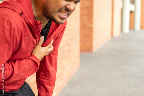 Sportsman wearing a red jacket leaning against a brick wall of clutching his chest acute pain possible heart attack. Hands male holding chest with symptom heart attack disease on city street.