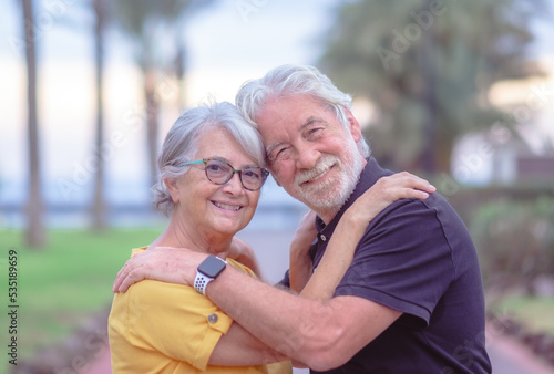Portrait of happy lovely senior couple embracing in outdoor public park at sunset. Two caucasian elderly people looking at camera smiling enjoying good time together