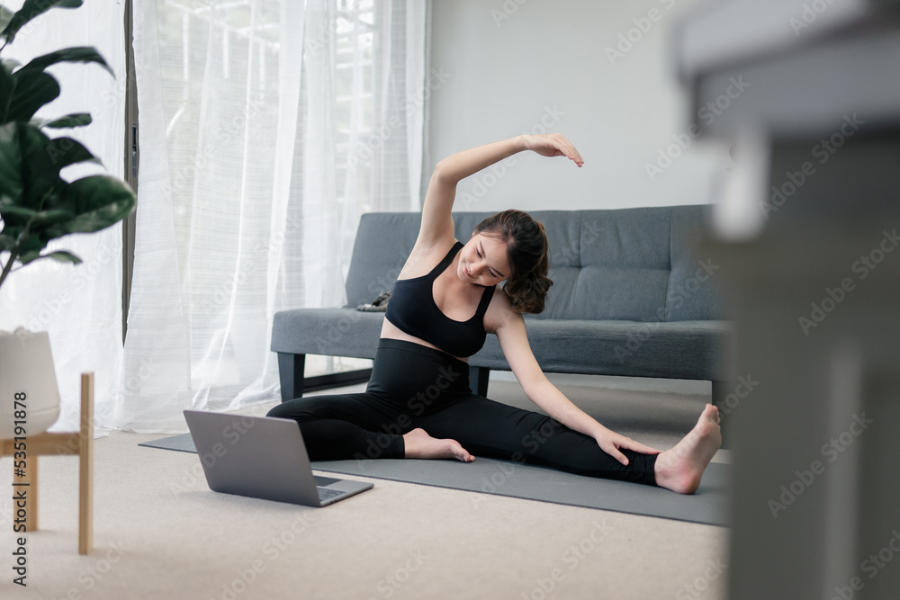 Beautiful pregnant asian woman is stretching her body in front of her laptop for the online yoga practicing to keep her prenatal healthy. Concept healthy pregnancy. 