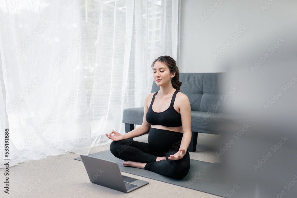 Beautiful pregnant asian woman is meditating in a lotus position in front of her laptop for the online yoga practicing in the living room to keep her prenatal healthy. Concept of Healthy Pregnancy. Beautiful pregnant asian woman is meditating in a lotus position in front of her laptop for the online yoga practicing in the living room to keep her prenatal healthy. Concept of Healthy Pregnancy.