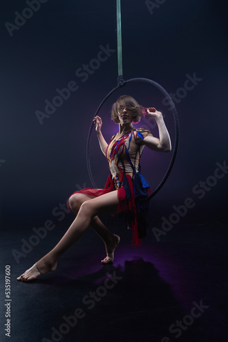 Beautiful woman performing acrobatic element on aerial ring indoors. Circus performer doing trick on arial lira on black background. © Naz