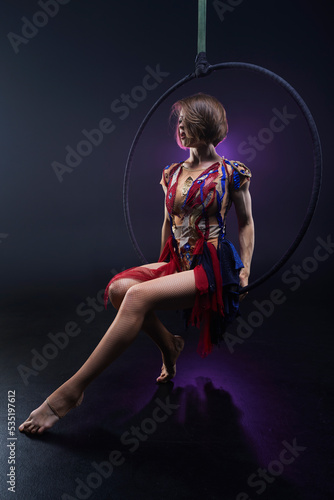 Beautiful woman performing acrobatic element on aerial ring indoors. Circus performer doing trick on arial lira on black background.