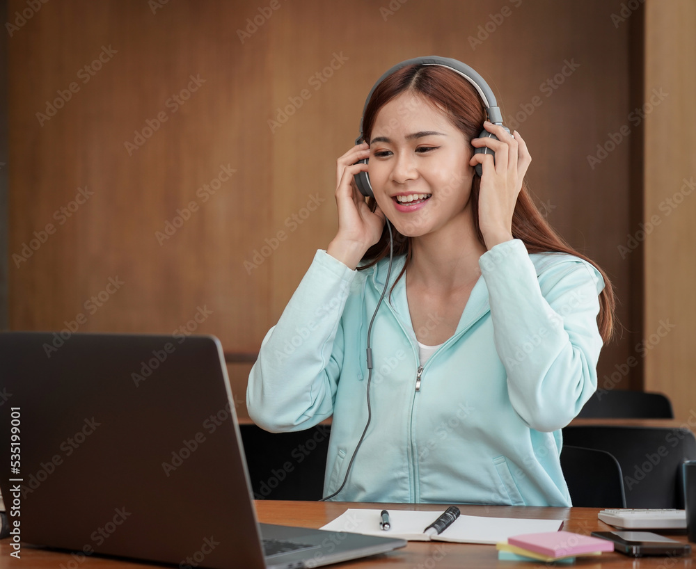 Female student is touching headphone and using laptop to studying online learning while taking notes Female student is touching headphone and using laptop to studying online learning while taking notes