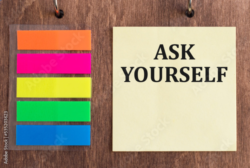 The words ASK YOURSELF written in a yellow card on a wooden background near multi-colored markers