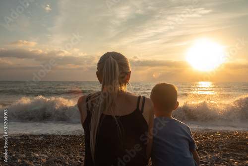 Mother and son are sitting on the beach near the sea and admiring beautifil sunset. family vacation concept