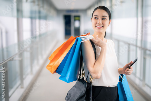 Asian woman holding smart phone with shopping bags walks through the city streets. Summer Style. Consumerism, purchases, shopping, lifestyle concept.