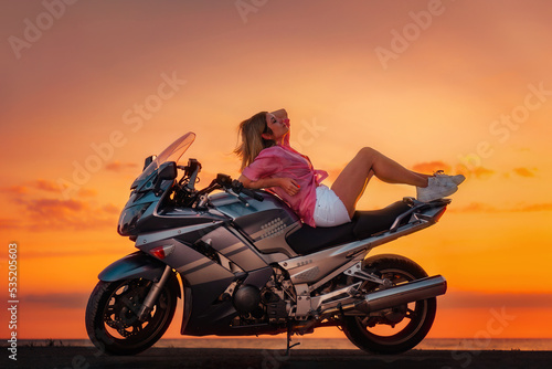 Sexy young woman posing on the motorcycle. Golden sunset and motorbike on the background. Copy space. Concept of World Motorcyclist Day and moto travel © _KUBE_